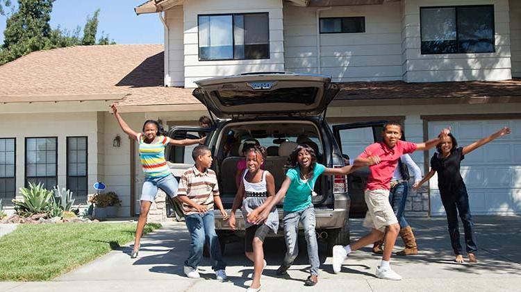 A gathering of young children frolic behind an open SUV parked in the driveway.